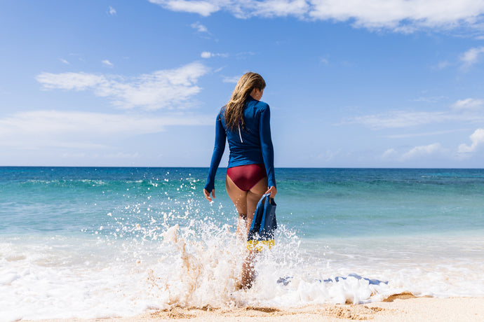 A woman in a blue long-sleeved rash guard and bikini bottoms walks towards the ocean on a sunny beach.